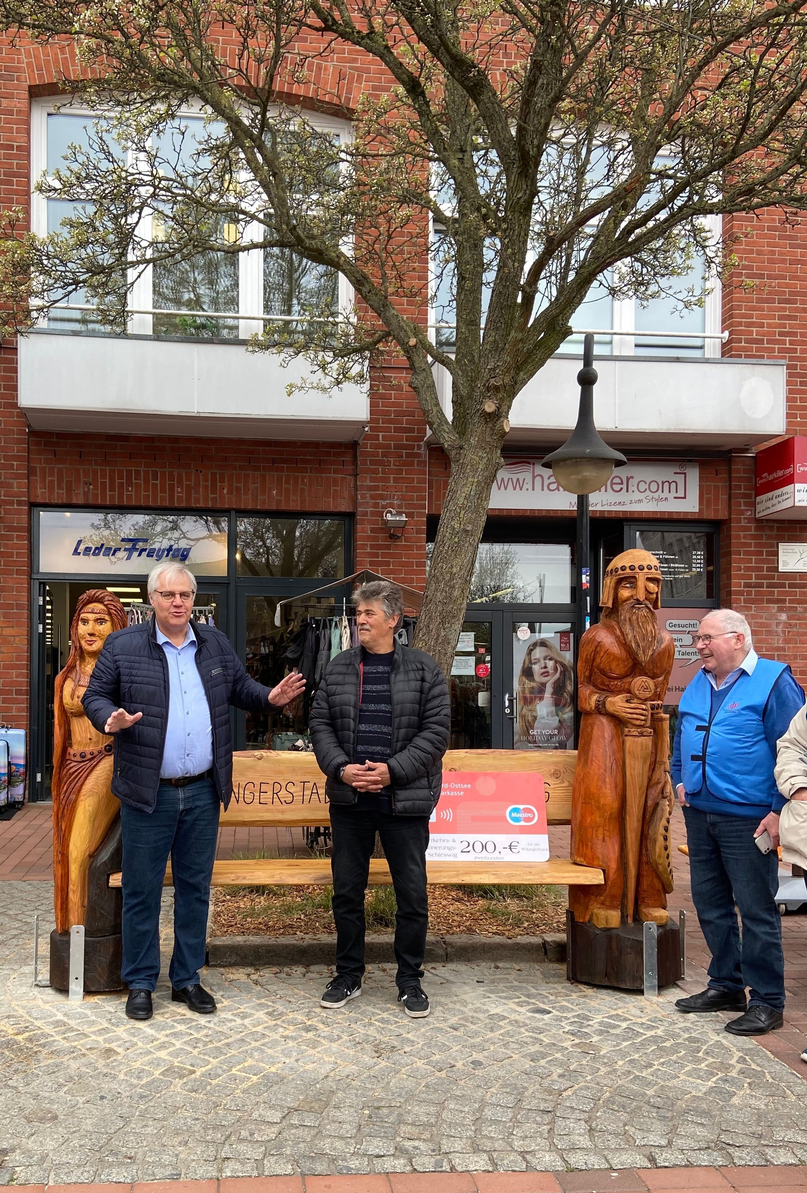 Wiederaufgebaute Wikingerbank am Capitolplatz in Schleswig, mit Sitzfläche aus Holz zwischen zwei Wikingerfiguren, symbolisch für die lokale Geschichte und Kultur.